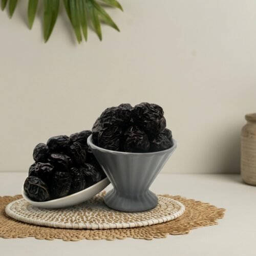 Premium Ajwa dates presented in a grey fluted pedestal bowl and side platter, highlighting the deep dark color and fine wrinkled texture of authentic Madinah dates.