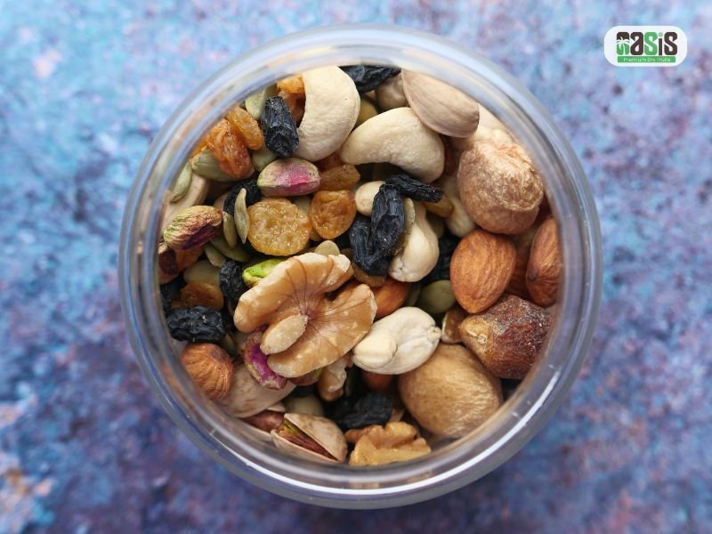 A top-down view of a glass jar filled with a healthy mix of dry fruits and nuts, including walnuts, almonds, and cashews, positioned against a blue textured background.