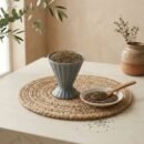 Premium organic ajwain carom seeds in a grey cup with wooden spoon on mat by Oasis Dry Fruits.