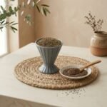 Premium organic ajwain carom seeds in a grey cup with wooden spoon on mat by Oasis Dry Fruits.