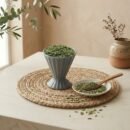 Green fennel seeds saunf in a grey cup with a wooden spoon on a mat by Oasis Dry Fruits.